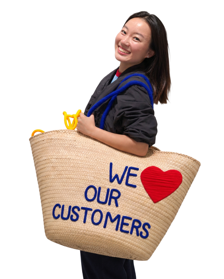 Sandy Zhao holding a woven tote bag that reads 'WE LOVE OUR CUSTOMERS'